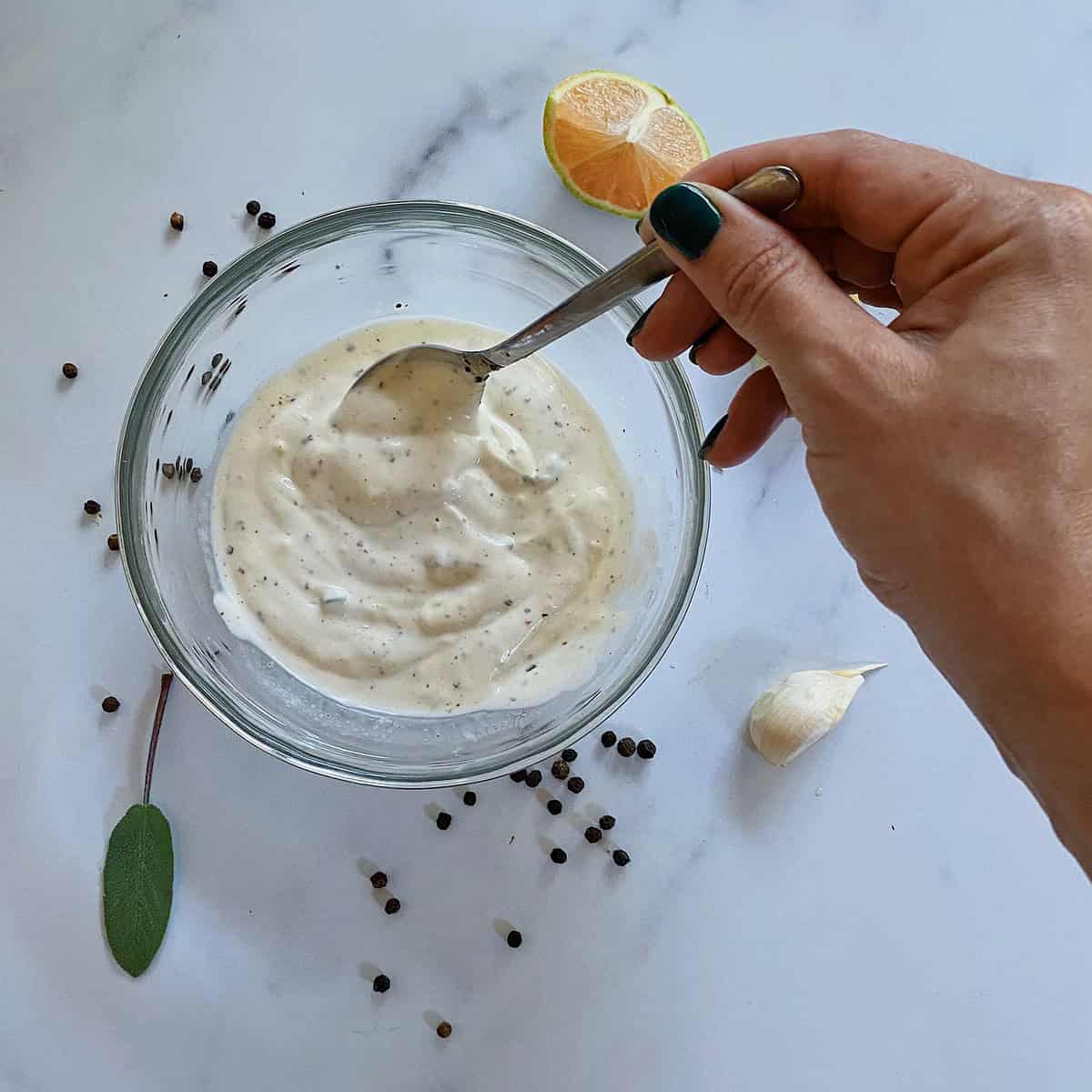 hand mixing aioli in clear bowl with black peppercorns, garlic and sage around it on a marble background