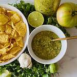 Bird's eye shot of fresh salsa verde with ingredients arrayed around the bowl and a stack of tortilla chips on the side