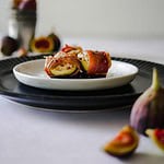 Warm baked figs wrapped in prosciutto and artistically photographed with a bucket of figs and a modern white background